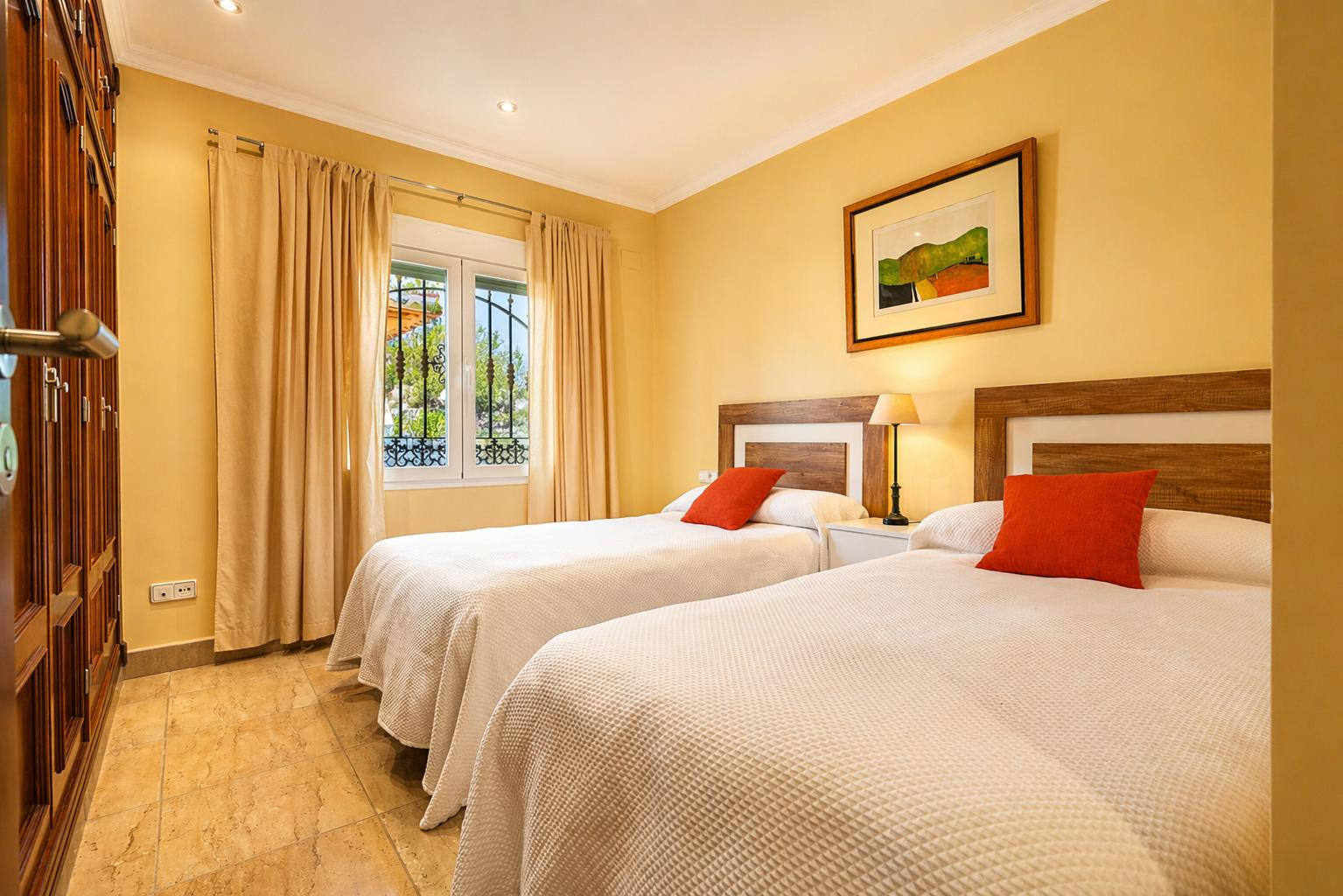 Dormitorio con dos camas individuales, cabeceros de madera y cojines naranjas. Dispone de armario empotrado, suelo de gres y ventana con rejas y cortinas beige.