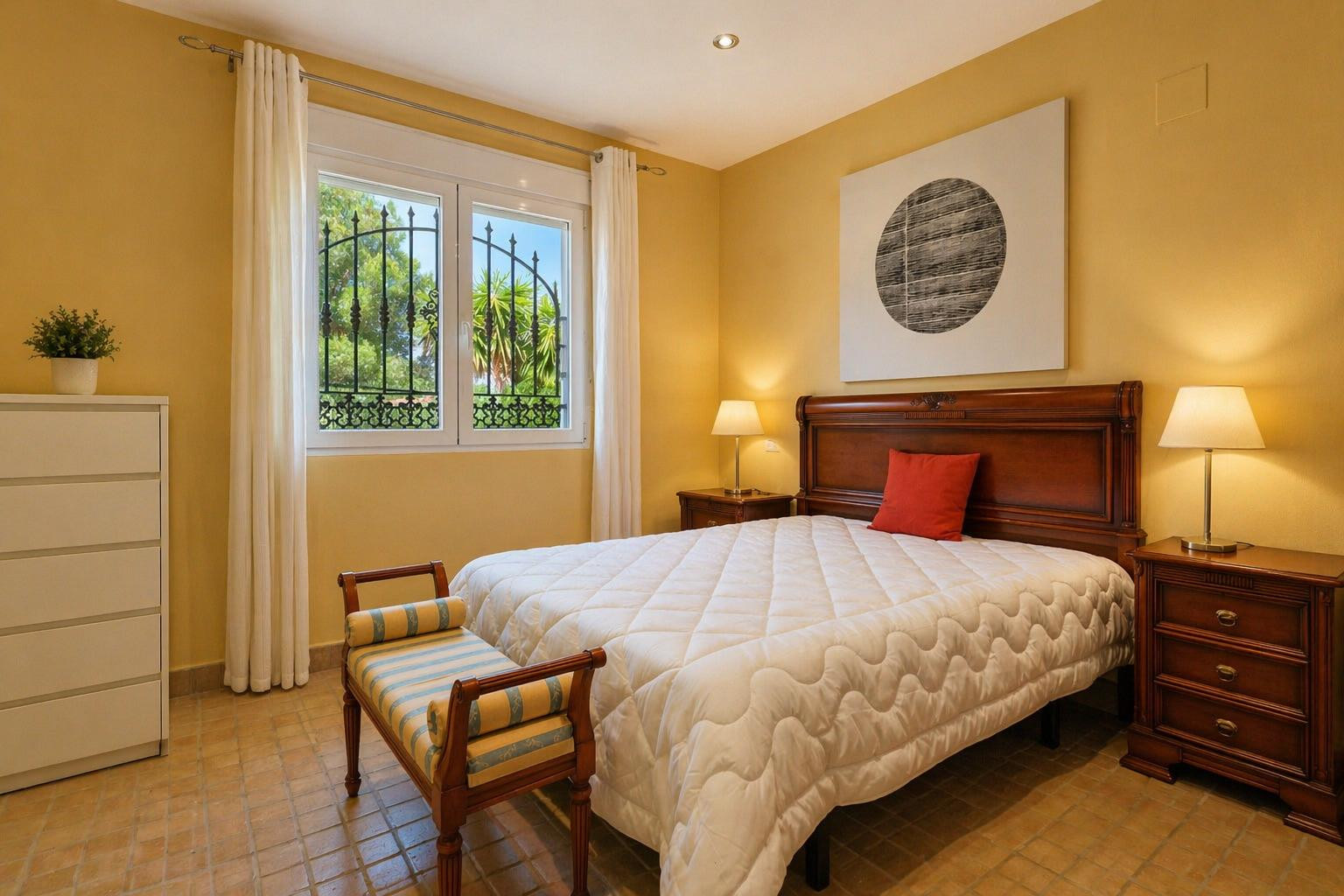 Dormitorio luminoso con cama de madera maciza, mesitas de noche a juego y suelo de terracota. La ventana con rejas de hierro forjado ofrece vistas al jardín.