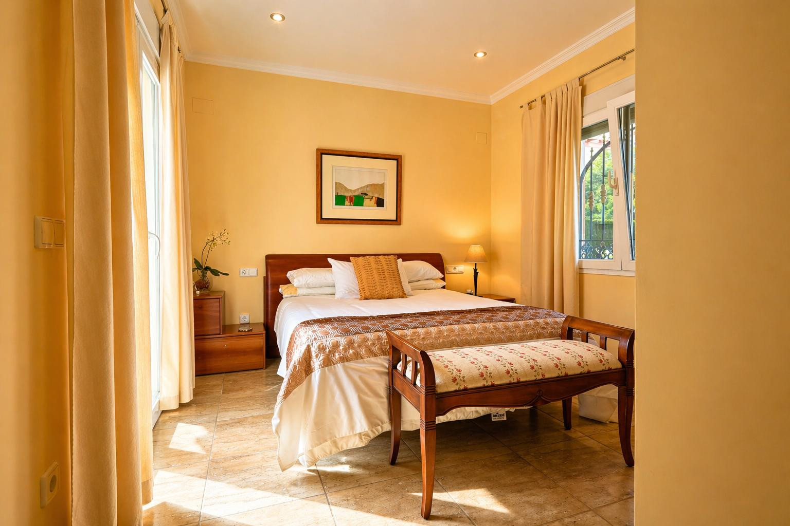 Dormitorio luminoso con cama doble, cabecero de madera y suelo de baldosas. Cuenta con acceso directo al exterior y una ventana con rejas decorativas.