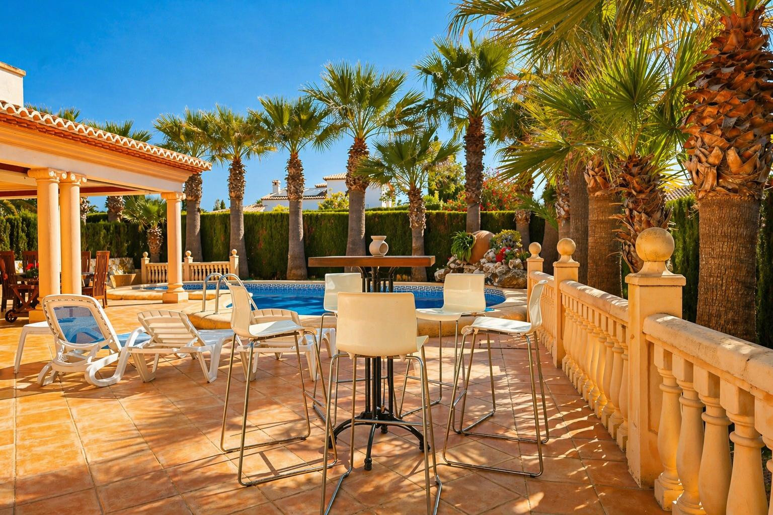 El patio exterior privado cuenta con una mesa alta y taburetes, tumbonas y una balaustrada, todo sobre baldosas de terracota. Una refrescante piscina está enmarcada por palmeras maduras y un seto frondoso.