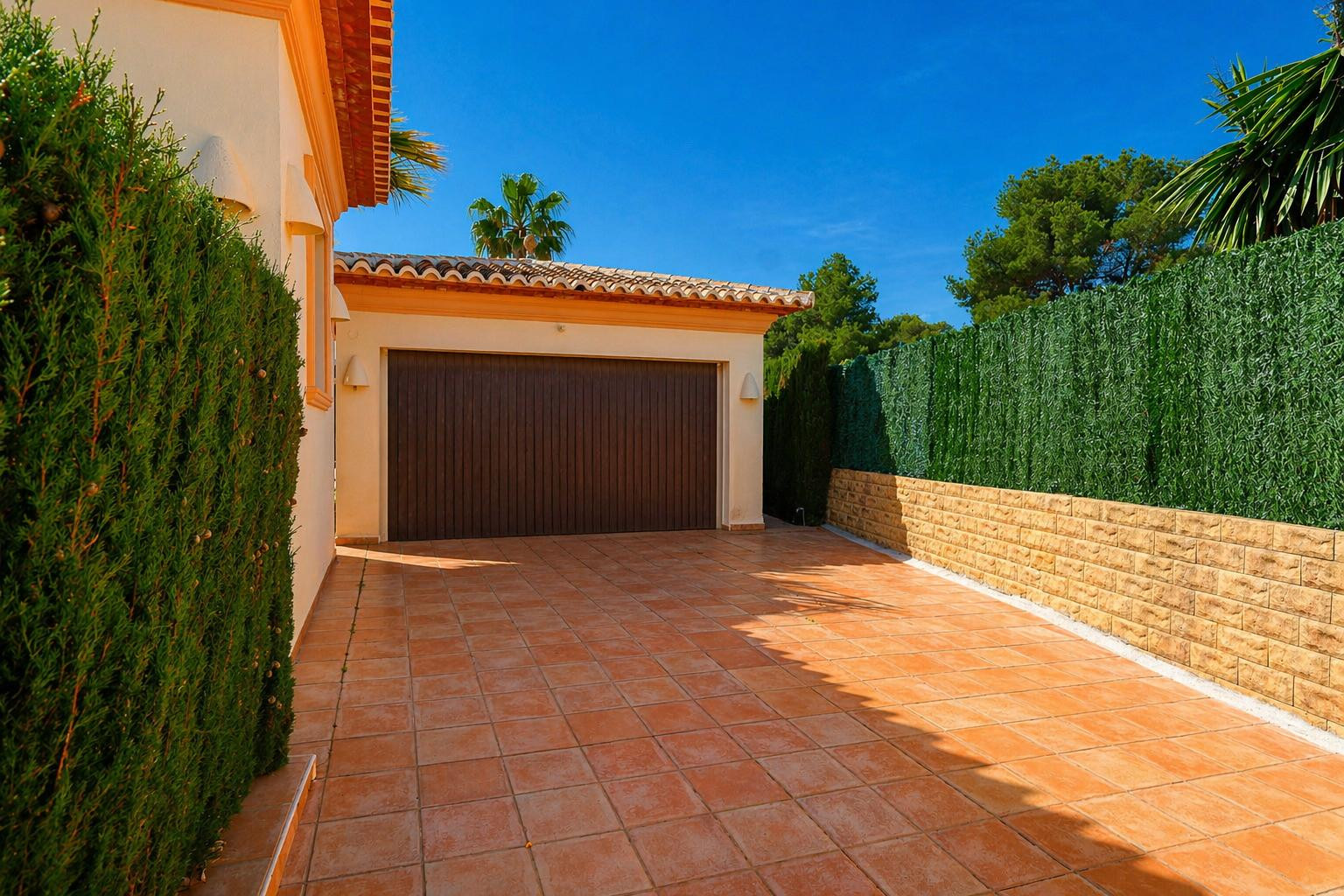 Garaje privado con puerta de madera oscura y entrada de baldosas de terracota, flanqueado por un seto natural y un muro de piedra con pantalla verde artificial, bajo un cielo azul claro.