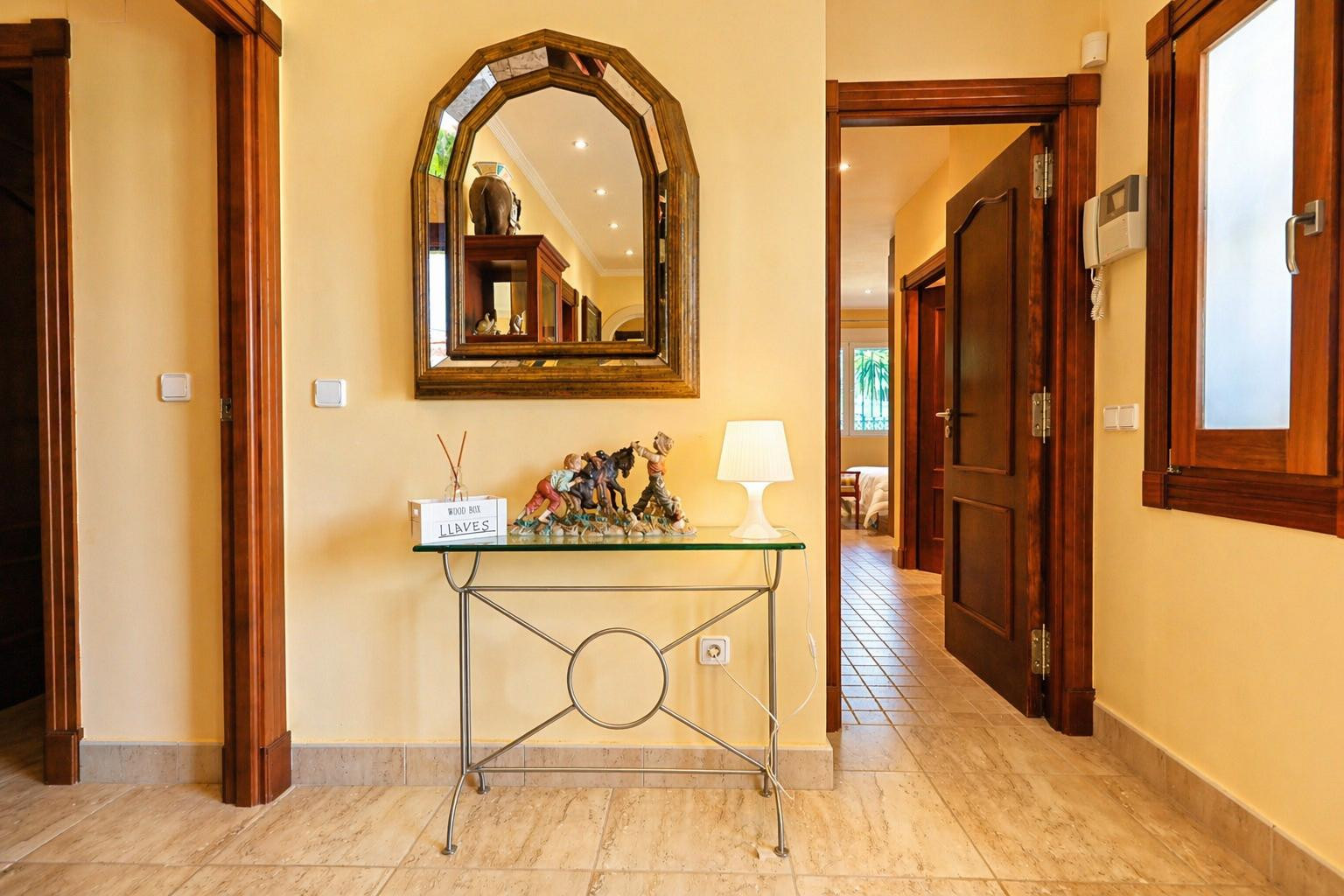 Hall de entrada luminoso con paredes amarillas y suelo de baldosas. Espejo arqueado de madera sobre consola de cristal, junto a puertas de madera oscura y un intercomunicador.