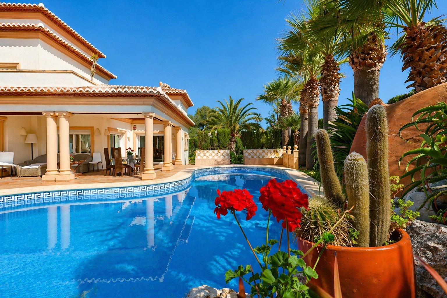 La atractiva piscina con su borde de mosaico de greca enmarca una amplia terraza cubierta, con elegantes zonas de comedor y salón exterior, rodeada de exuberantes palmeras y cactus mediterráneos.