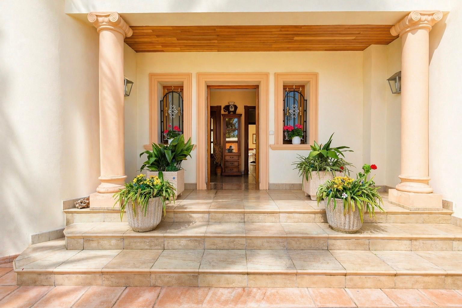 La elegante entrada principal presenta columnas clásicas con capiteles jónicos, escalones de piedra y un cálido techo de madera. Plantas en maceta adornan el porche, flanqueando la puerta abierta y ventanas con rejas.