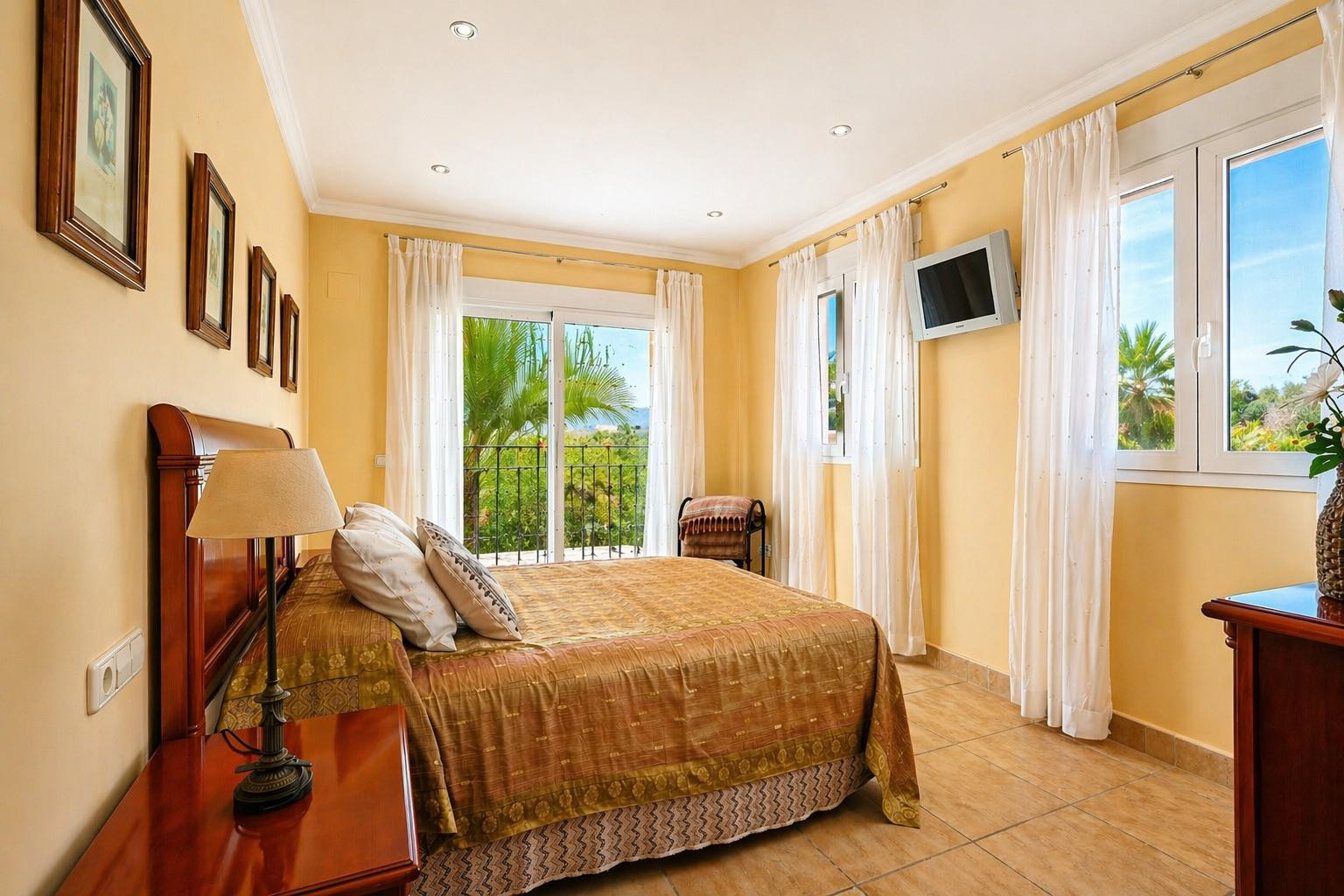 Luminoso dormitorio con cama doble y cabecero de madera, suelo de baldosas y acceso directo a un balcón privado con vistas a palmeras y vegetación.