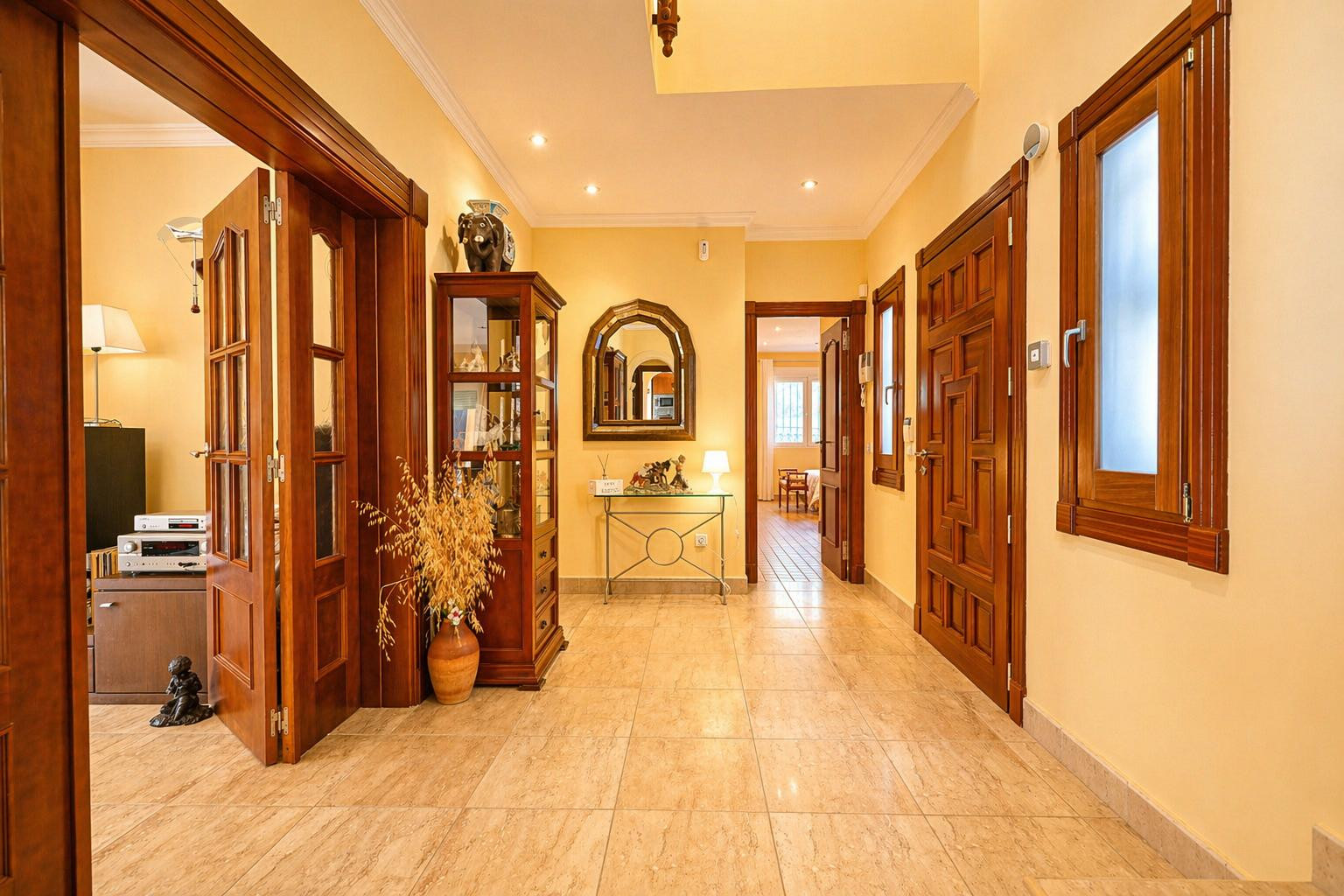 Luminoso pasillo con paredes amarillas cálidas y suelo de baldosas claras. Destacan las puertas de madera oscura, una vitrina decorativa y una consola con espejo arqueado.