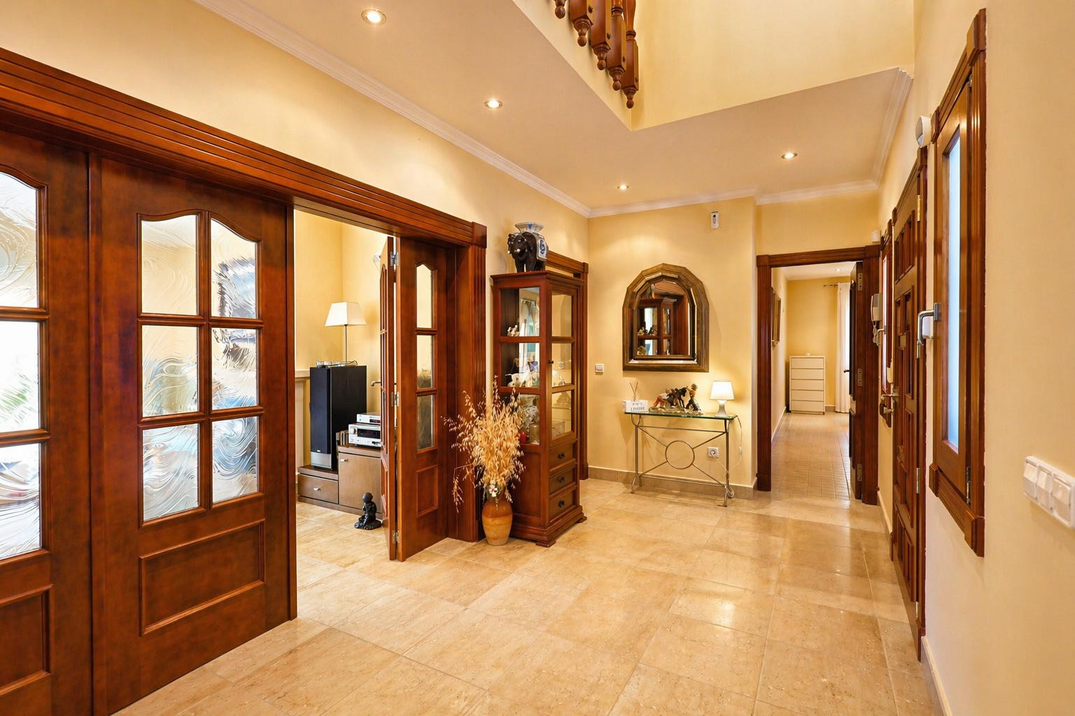 Luminoso pasillo con suelo de baldosas claras y focos empotrados. Puertas de madera oscura con paneles de cristal decorado conectan las estancias, junto a una consola y vitrina.