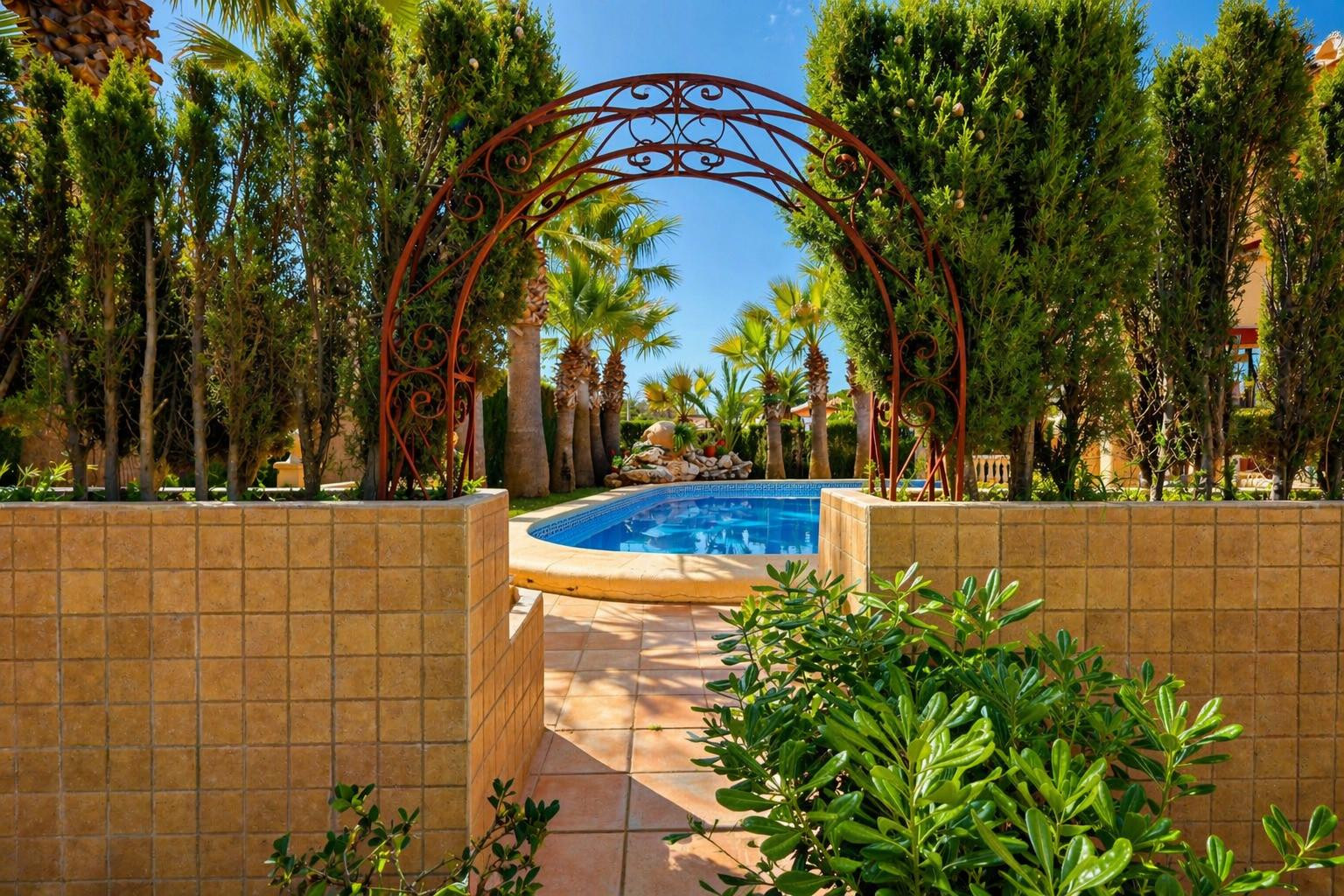 Un arco de hierro forjado ornamentado enmarca un camino de baldosas que conduce a la atractiva piscina azul, rodeada de exuberante vegetación y palmeras.