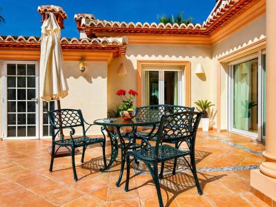 Amplia terraza exterior con suelo de terracota y un borde de azulejos decorativos. Destaca un conjunto de comedor de metal verde oscuro y elementos arquitectónicos mediterráneos.