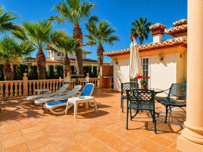 Amplia terraza exterior pavimentada con baldosas de terracota, equipada con tumbonas y un conjunto de mesa y sillas de metal bajo una sombrilla. Palmeras y una balaustrada de piedra enmarcan el espacio.