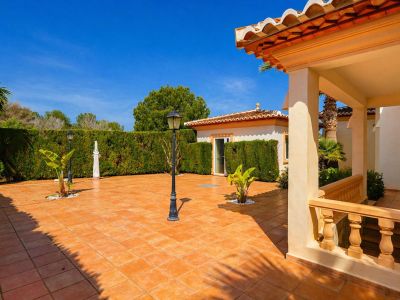 Amplio patio exterior con baldosas de terracota, delimitado por un seto verde, que incluye farolas clásicas y un porche cubierto con balaustrada y tejado tradicional.