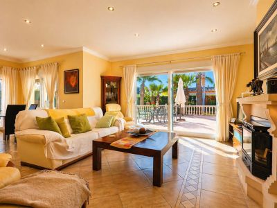 Amplio salón-comedor con suelo de baldosas y chimenea, que se abre a la terraza exterior con palmeras a través de grandes puertas correderas de cristal.