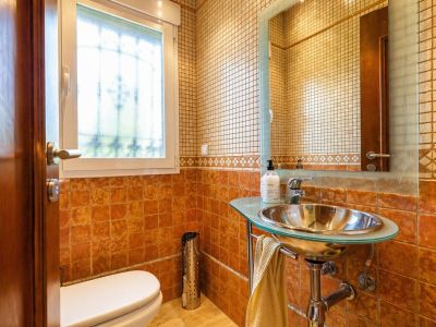 Aseo de cortesía con azulejos terracota y mosaico, lavabo moderno con encimera de cristal y bol metálico, y ventana que aporta luz natural.