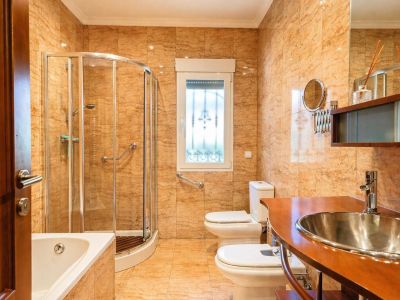 Baño completo con azulejos de tonos tierra, bañera, ducha de cristal curvada con barras de apoyo, inodoro, bidé y lavabo de acero inoxidable sobre mueble de madera.