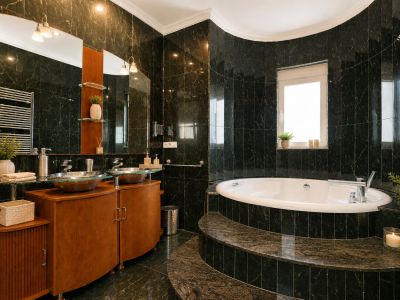 Baño elegante con azulejos de patrón mármol verde oscuro, bañera curva integrada sobre escalones de piedra y doble lavabo de cristal sobre mueble de madera.