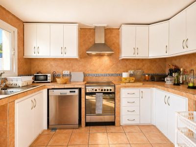 Cocina funcional equipada con azulejos de terracota en suelo y pared, armarios blancos y electrodomésticos de acero inoxidable. La luz natural entra por la ventana sobre el fregadero.