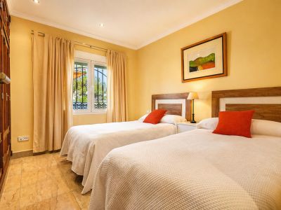 Dormitorio con dos camas individuales, cabeceros de madera y cojines naranjas. Dispone de armario empotrado, suelo de gres y ventana con rejas y cortinas beige.