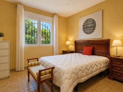 Dormitorio luminoso con cama de madera maciza, mesitas de noche a juego y suelo de terracota. La ventana con rejas de hierro forjado ofrece vistas al jardín.