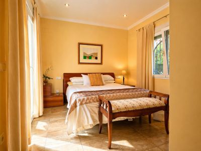 Dormitorio luminoso con cama doble, cabecero de madera y suelo de baldosas. Cuenta con acceso directo al exterior y una ventana con rejas decorativas.