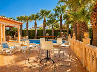 El patio exterior privado cuenta con una mesa alta y taburetes, tumbonas y una balaustrada, todo sobre baldosas de terracota. Una refrescante piscina está enmarcada por palmeras maduras y un seto frondoso.