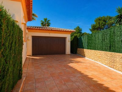 Garaje privado con puerta de madera oscura y entrada de baldosas de terracota, flanqueado por un seto natural y un muro de piedra con pantalla verde artificial, bajo un cielo azul claro.