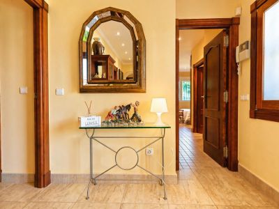 Hall de entrada luminoso con paredes amarillas y suelo de baldosas. Espejo arqueado de madera sobre consola de cristal, junto a puertas de madera oscura y un intercomunicador.
