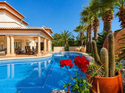 La atractiva piscina con su borde de mosaico de greca enmarca una amplia terraza cubierta, con elegantes zonas de comedor y salón exterior, rodeada de exuberantes palmeras y cactus mediterráneos.