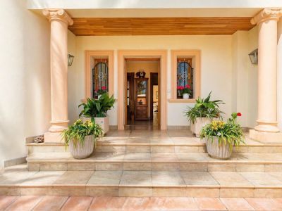 La elegante entrada principal presenta columnas clásicas con capiteles jónicos, escalones de piedra y un cálido techo de madera. Plantas en maceta adornan el porche, flanqueando la puerta abierta y ventanas con rejas.