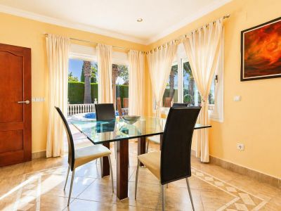 Luminoso comedor con mesa de cristal, sillas modernas y suelo de baldosas con patrón geométrico. Grandes ventanales ofrecen vistas directas al jardín y la piscina privada.