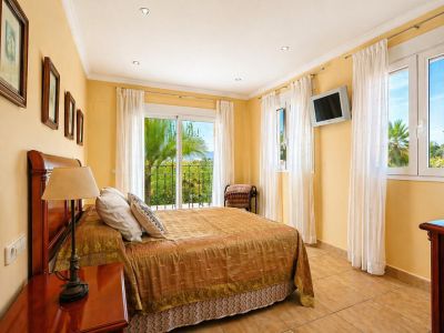 Luminoso dormitorio con cama doble y cabecero de madera, suelo de baldosas y acceso directo a un balcón privado con vistas a palmeras y vegetación.