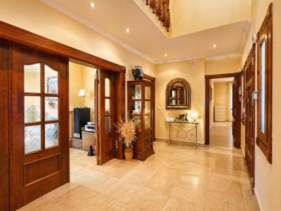 Luminoso pasillo con suelo de baldosas claras y focos empotrados. Puertas de madera oscura con paneles de cristal decorado conectan las estancias, junto a una consola y vitrina.