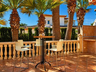 Terraza exterior con suelo de baldosas de terracota, balustrada clásica y un conjunto de mesa alta con taburetes. Incluye una zona de barbacoa integrada y palmeras que aportan sombra.