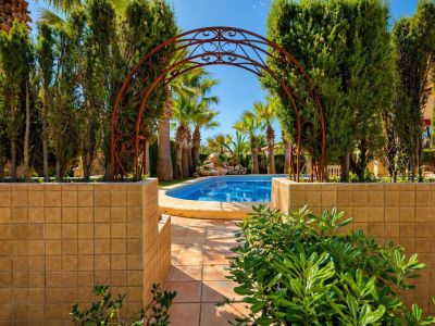 Un arco de hierro forjado ornamentado enmarca un camino de baldosas que conduce a la atractiva piscina azul, rodeada de exuberante vegetación y palmeras.