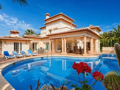 Vista exterior de la villa con piscina curva de mosaico azul y solárium. La terraza cubierta con columnas alberga zonas de estar y comedor al aire libre, conectando directamente con el jardín.