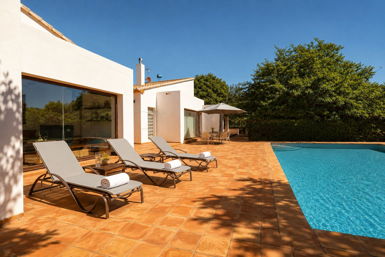 Amplia terraza de gres rústico con piscina, rodeada de tres tumbonas y una zona de comedor exterior bajo sombrilla. La fachada blanca con grandes ventanales refleja el entorno natural. Amplia terraza de gres rústico con piscina, rodeada de tres tumbonas y una zona de comedor exterior bajo sombrilla. La fachada blanca con grandes ventanales refleja el entorno natural.