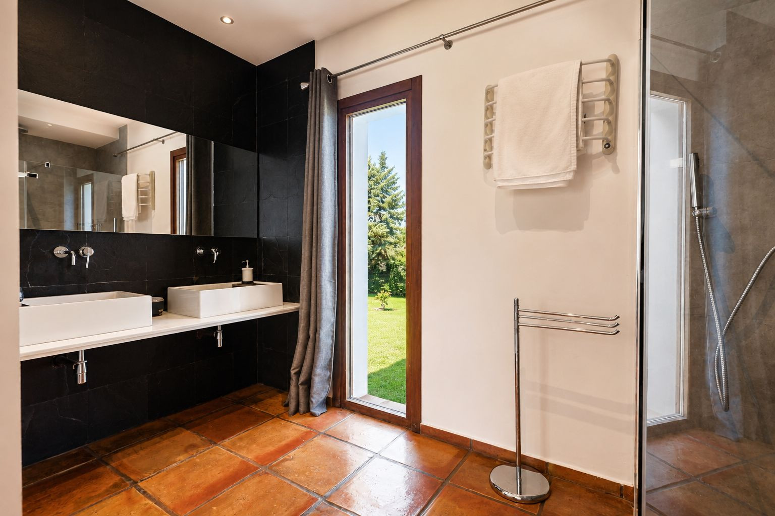 Amplio baño con suelos de barro, doble lavabo rectangular blanco sobre encimera flotante y pared de azulejos negros. Gran ventanal con vistas al jardín y ducha acristalada. Amplio baño con suelos de barro, doble lavabo rectangular blanco sobre encimera flotante y pared de azulejos negros. Gran ventanal con vistas al jardín y ducha acristalada.