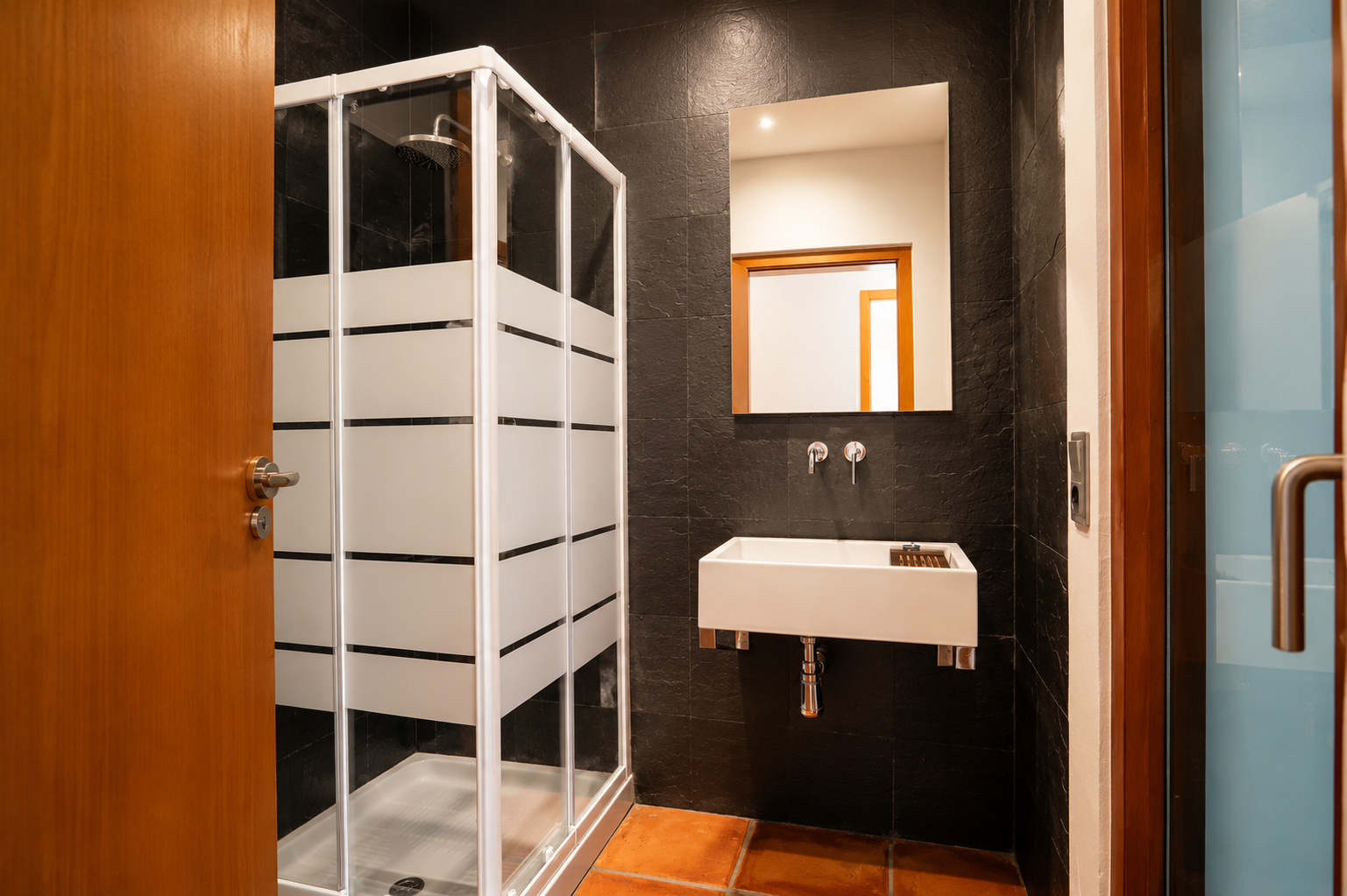 Baño con ducha acristalada de franjas esmeriladas, lavabo rectangular suspendido y grifería mural. Paredes de azulejos oscuros texturizados y suelo de barro cocido. Baño con ducha acristalada de franjas esmeriladas, lavabo rectangular suspendido y grifería mural. Paredes de azulejos oscuros texturizados y suelo de barro cocido.
