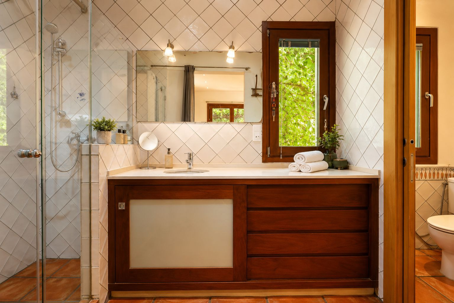 Baño con ducha acristalada, tocador de madera oscura y encimera blanca. Ventana con vistas a la vegetación, azulejos blancos en rombo y suelos de barro artesanal. Baño con ducha acristalada, tocador de madera oscura y encimera blanca. Ventana con vistas a la vegetación, azulejos blancos en rombo y suelos de barro artesanal.