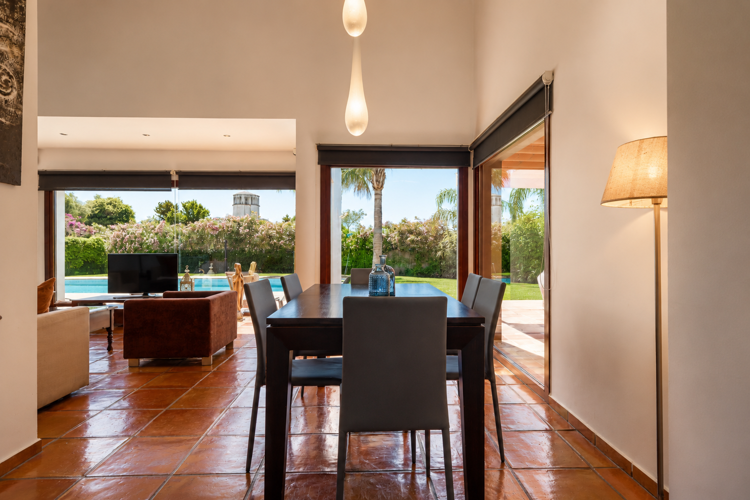 Comedor con mesa de madera oscura y suelos de barro artesanal, bañados por luz natural. Las grandes puertas de cristal ofrecen vistas directas al jardín, piscina y una torre lejana. Comedor con mesa de madera oscura y suelos de barro artesanal, bañados por luz natural. Las grandes puertas de cristal ofrecen vistas directas al jardín, piscina y una torre lejana.