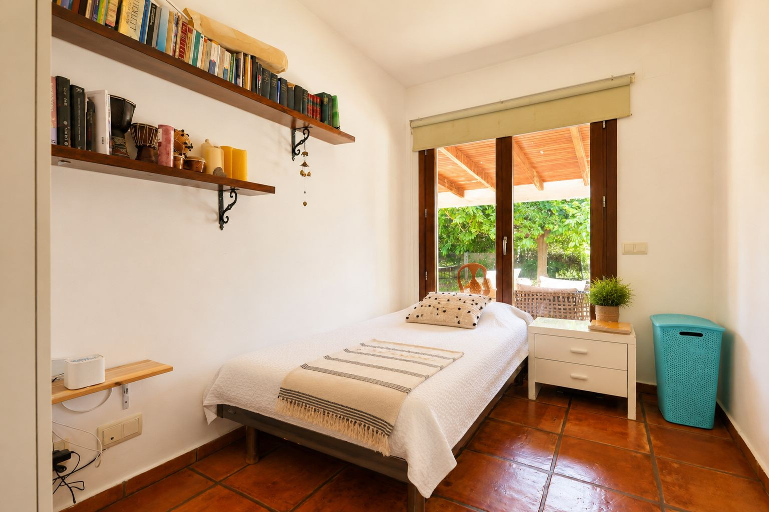Dormitorio individual con suelos de barro artesanal, paredes blancas y acceso directo a una terraza cubierta a través de una gran puerta de cristal con marco de madera. Incluye estantes con libros. Dormitorio individual con suelos de barro artesanal, paredes blancas y acceso directo a una terraza cubierta a través de una gran puerta de cristal con marco de madera. Incluye estantes con libros.