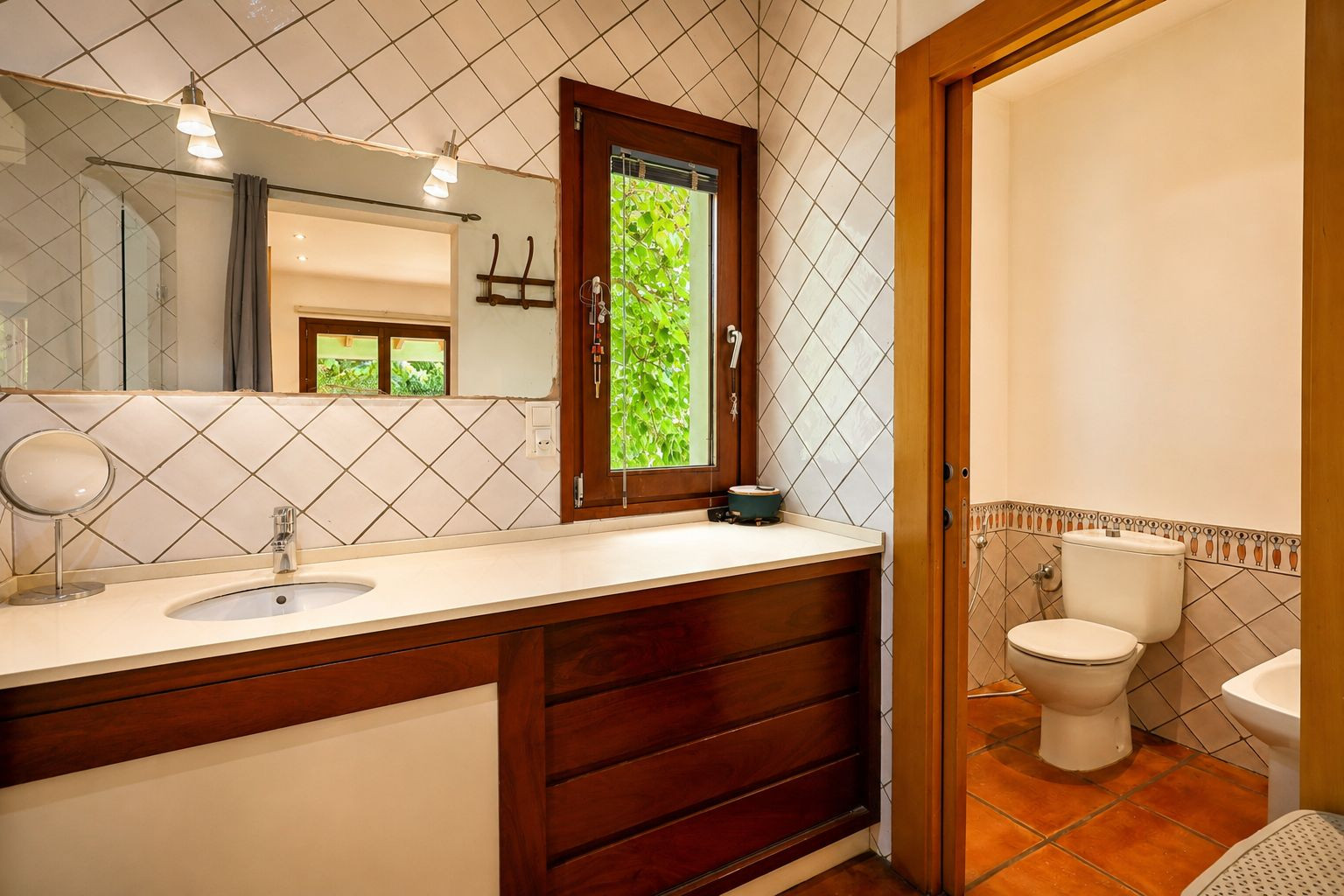 Lavabo con encimera clara y mueble de madera oscura. El gran espejo con apliques ilumina la zona, mientras una ventana de madera ofrece vistas a la vegetación exterior. Lavabo con encimera clara y mueble de madera oscura. El gran espejo con apliques ilumina la zona, mientras una ventana de madera ofrece vistas a la vegetación exterior.