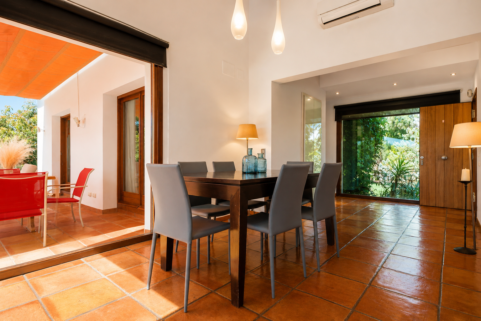 Luminoso comedor con mesa de madera oscura y seis sillas grises, sobre suelos de barro artesanal. Grandes puertas de cristal conectan con una terraza exterior bajo toldo naranja y vistas al jardín. Luminoso comedor con mesa de madera oscura y seis sillas grises, sobre suelos de barro artesanal. Grandes puertas de cristal conectan con una terraza exterior bajo toldo naranja y vistas al jardín.