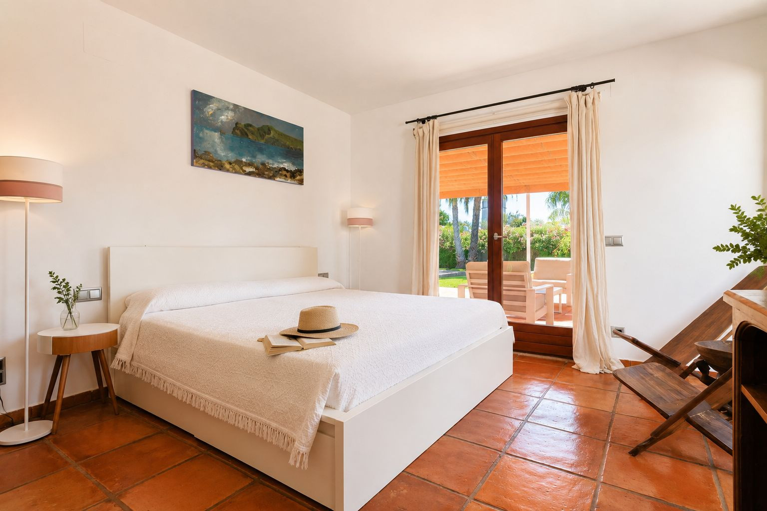 Luminoso dormitorio con cama blanca, suelos de barro artesanal y acceso directo a una terraza exterior con vistas al jardín y mobiliario. Luminoso dormitorio con cama blanca, suelos de barro artesanal y acceso directo a una terraza exterior con vistas al jardín y mobiliario.
