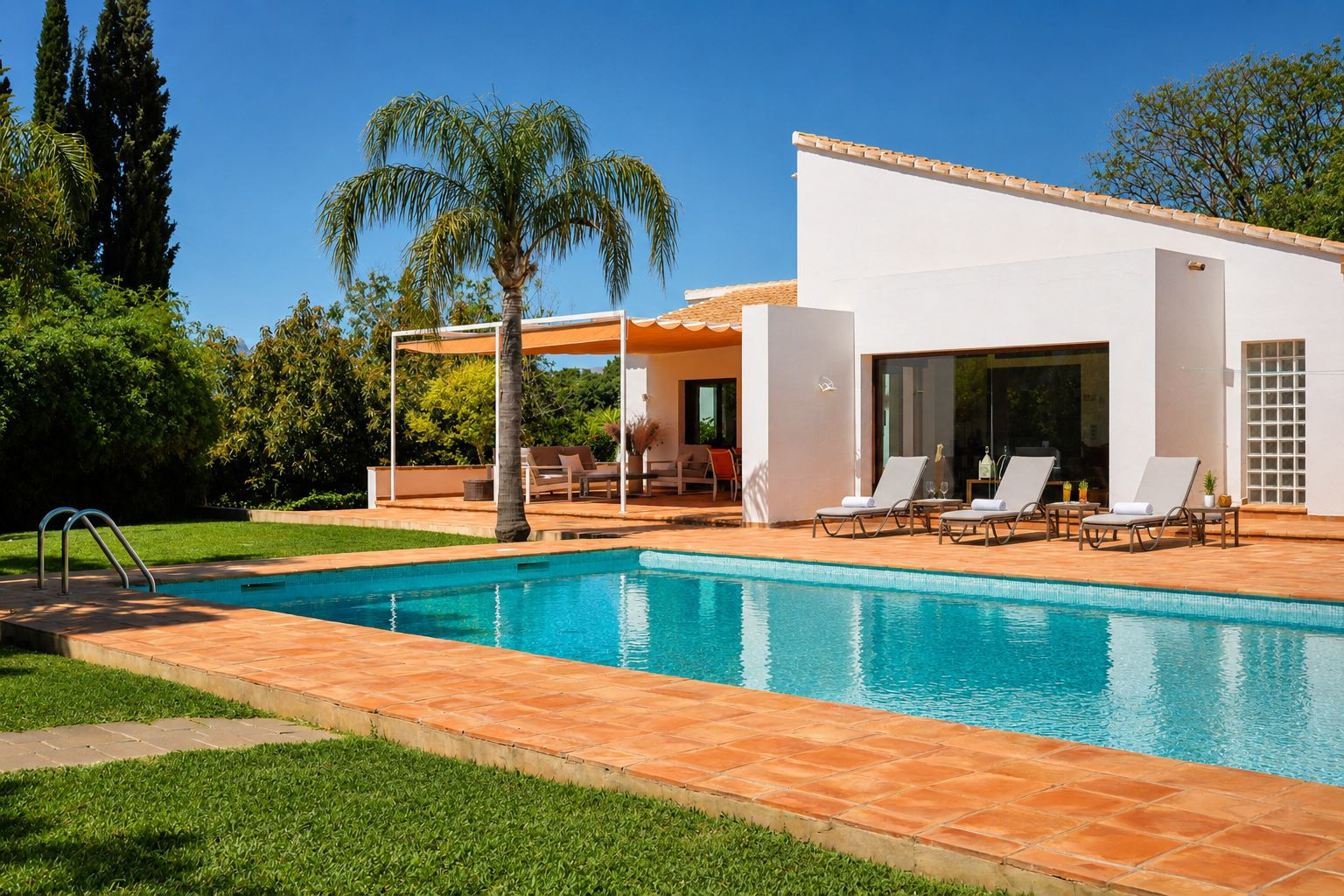 Piscina de azulejos azules rodeada de césped y baldosas de barro, con tumbonas junto a la fachada blanca de la vivienda principal y una pérgola sombreada con zona lounge. Piscina de azulejos azules rodeada de césped y baldosas de barro, con tumbonas junto a la fachada blanca de la vivienda principal y una pérgola sombreada con zona lounge.