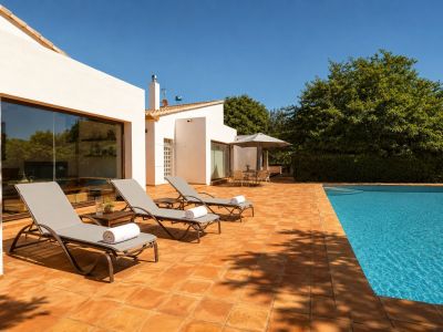 Amplia terraza de gres rústico con piscina, rodeada de tres tumbonas y una zona de comedor exterior bajo sombrilla. La fachada blanca con grandes ventanales refleja el entorno natural.