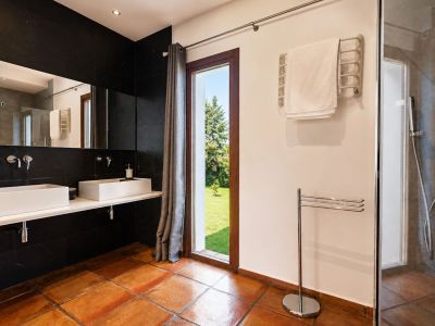 Amplio baño con suelos de barro, doble lavabo rectangular blanco sobre encimera flotante y pared de azulejos negros. Gran ventanal con vistas al jardín y ducha acristalada.