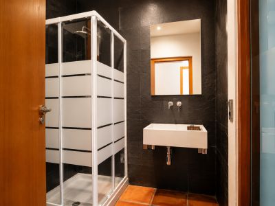 Baño con ducha acristalada de franjas esmeriladas, lavabo rectangular suspendido y grifería mural. Paredes de azulejos oscuros texturizados y suelo de barro cocido.