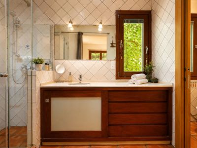 Baño con ducha acristalada, tocador de madera oscura y encimera blanca. Ventana con vistas a la vegetación, azulejos blancos en rombo y suelos de barro artesanal.
