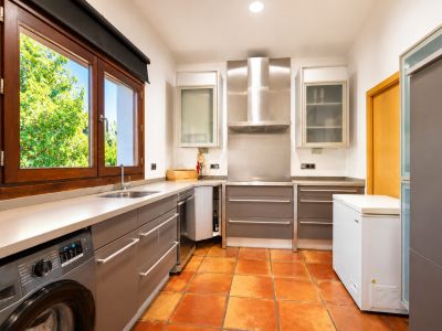 Cocina funcional con lavadora integrada, suelo de barro artesanal y gran ventanal de madera con vistas al jardín.