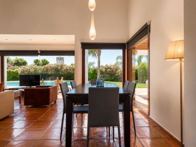 Comedor con mesa de madera oscura y suelos de barro artesanal, bañados por luz natural. Las grandes puertas de cristal ofrecen vistas directas al jardín, piscina y una torre lejana.